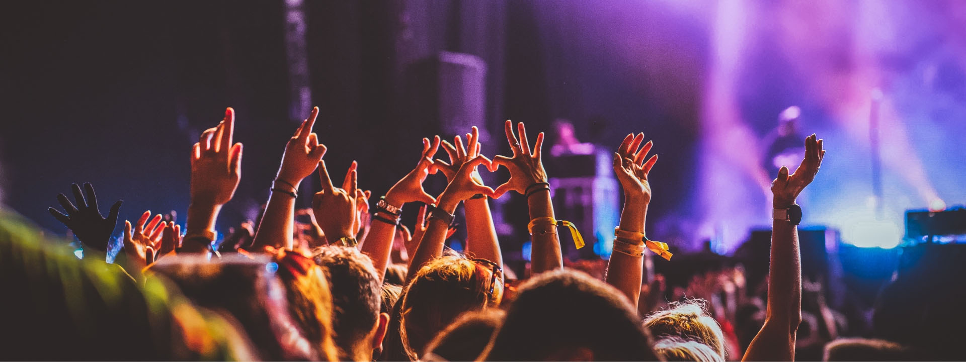 A crowd at a concert raising their hands, some forming heart shapes, against a backdrop of colorful stage lighting.