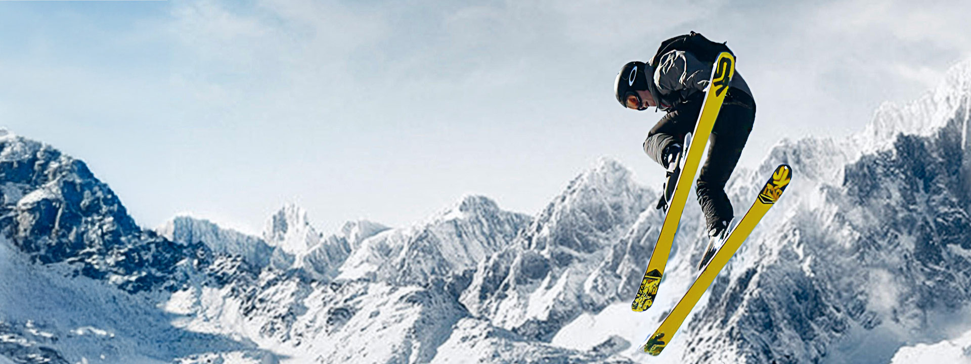 A skier in mid-air performs a trick against a snowy mountain backdrop, holding yellow skis with a clear sky in the background.