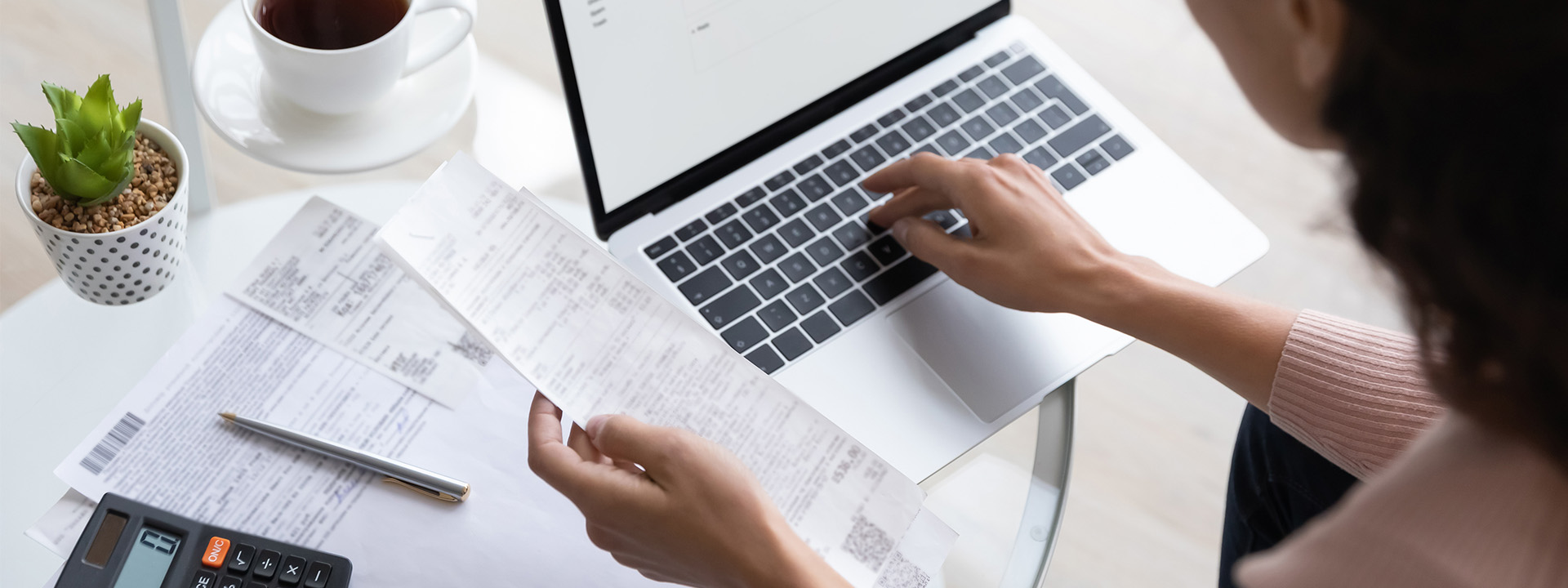 A person is working on a laptop at a table, holding a receipt in one hand and typing with the other. Papers, a calculator, a pen, and a small plant are also on the table.