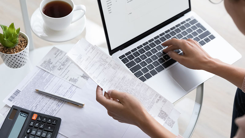 A person is working on a laptop at a table, holding a receipt in one hand and typing with the other. Papers, a calculator, a pen, and a small plant are also on the table.