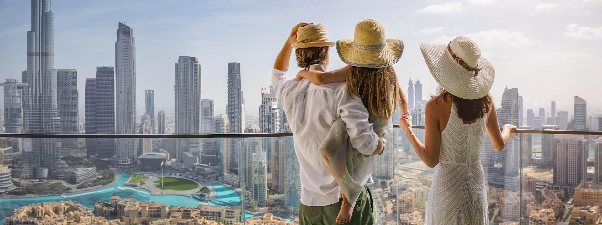 A family of three wearing hats stands on a balcony overlooking a city with tall buildings and a distant view of a body of water.