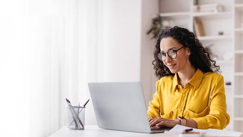 A woman in a yellow blouse and glasses smiling while working on a laptop in a bright office space.